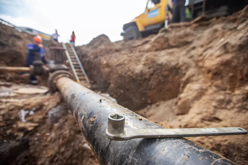 Close-up of exposed pipe in trench with blurred utility workers and yellow truck in background.